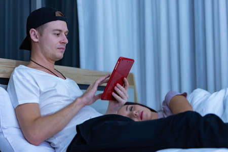 Man sitting on bed in the night and using tablet while wife sleeping beside him.の写真素材