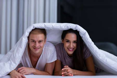 Portrait of young happy playful couple relaxing in comfortable cozy bed, cheerful smiling man and woman having fun posing covering head with soft warm blanket duvet on sleepoverの写真素材