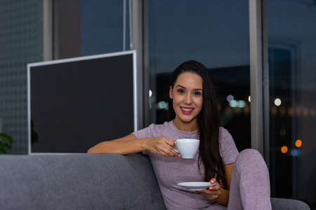 Woman enjoying a cup of tea in the night sitting on a couch in the living room at homeの写真素材