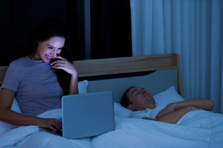 Woman sitting on bed in the night and using laptop computer while husband sleeping beside her.の写真素材
