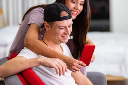 Happy couple enjoying media content in a smart phone sitting on a couch at homeの写真素材