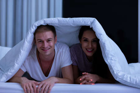 Portrait of young happy playful couple relaxing in comfortable cozy bed, cheerful smiling man and woman having fun posing covering head with soft warm blanket duvet on sleepoverの写真素材