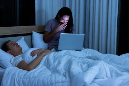 Woman sitting on bed in the night and using laptop computer while husband sleeping beside her.の写真素材