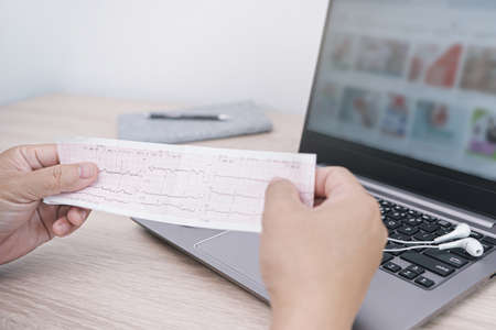 Close up Heart rate result graph paper document electrocardiogram  in hand a man analysis of medical research results and laptop computer in backgroundの写真素材