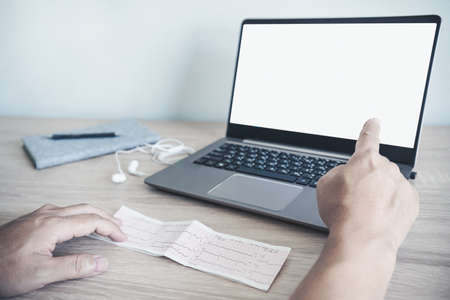 Close up Heart rate result graph paper document electrocardiogram on wood table and a man use smartphone and laptop computer Search about healthの写真素材