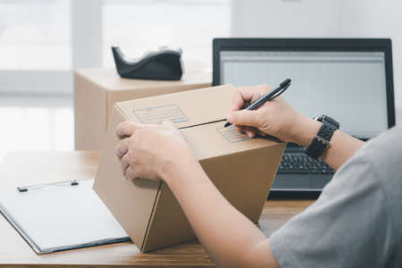 Close up hand a man use pen write on parcel box about the parcel delivery address and use laptop to...