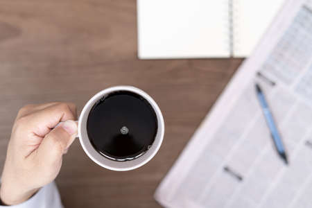 A man hold coffee cup drink black coffee and pen newspaper notebook on vintage wood tableの写真素材