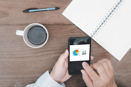 Close hand a man hold smartphone open work for check and pen notebook coffee cup on table