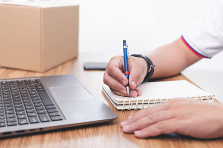 Parcel delivery staff use pencil write on notebook paper About delivery information of customers and use laptop smartphone Parcel box on the tableの写真素材