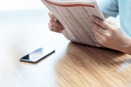 Close up hand a man hold smartphone reading news online and newspaper ice coffee on wood table relax time in holidayの写真素材