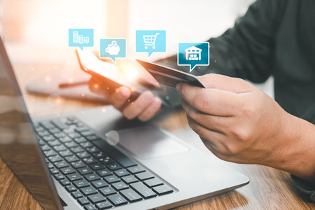 Man wearing a green shirt holding a credit card and using a smartphone for payment online on mobile banking with a laptop on the table in a homeの写真素材