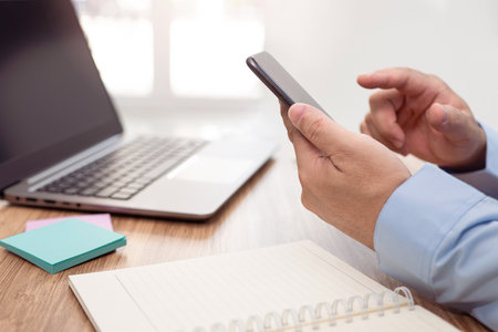 Businessman hold smartphone with finger touch to screen display and notebook and laptop on the table and working in office and glass window in blur backgroundの写真素材