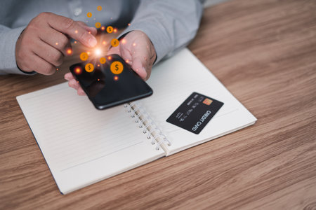 A man holding a smartphone pays money mobile banking online a credit card on a notebook with wood table in a home, Shopping online concept, and a money icon on the displayの写真素材