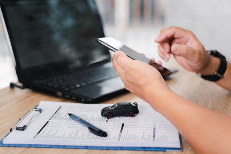 Car insurance online concept : A man holds a smartphone with checklist, toy car, Pen on table and use laptop check online case of accidentの写真素材