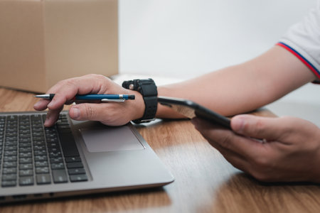 Close up hand a man parcel delivery staff typing on laptop keyboard and hold smartphone check parcel delivery information online data for accuracy before sending products to customの写真素材