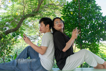 Two Thai LGBTQ friends relaxing back to back while playing mobile games together in the afternoon showing friendship relaxation connection and joyful lifestyleの写真素材