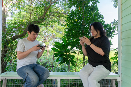 Two Thai LGBTQ friends playing mobile games together on a relaxing afternoon at home surrounded by...
