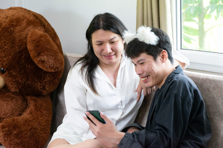 Two Thai LGBTQ friends smiling while checking social media together on smartphone showing happiness diversity friendship and modern lifestyle at homeの写真素材
