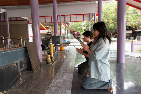 Two LGBTQ friends kneeling and praying at a Thai temple. One is holding flowers, while the other is in a peaceful prayer posture. They are performing a spiritual ritual in a tranquil temple setting.の写真素材