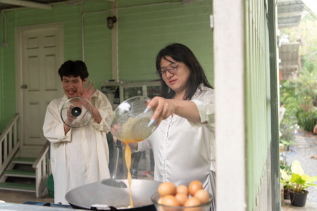 LGBTQ friends, one with long hair and one with short, share a joyful moment preparing a Thai-style omelette for breakfast in a cozy open kitchen. The scene captures the fun of cooking together.の写真素材