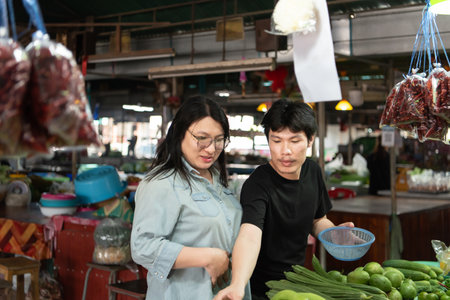 Two LGBTQ friends enjoying a shopping trip at a fresh market in Thailand. One picks vegetables, while the other smiles, sharing a joyful moment in the lively, colorful market atmosphere.の写真素材
