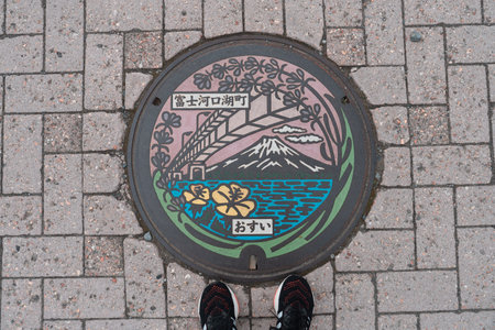 KAWAGUCHIKO, JAPAN - APRIL 7, 2023: Top view of manhole cover in Kawaguchiko, Japan with feet standing nearbyのeditorial素材