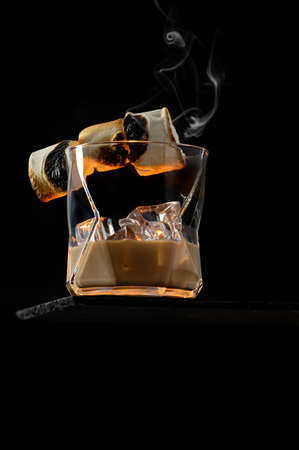 A glass of Irish Cream Liquor with ice cubes and garnished with burning caramelised marshmallow on black stone with low point of viewの写真素材
