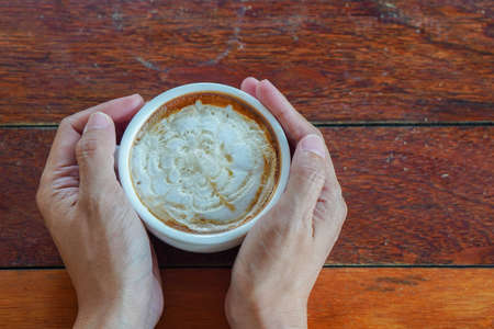 left side photo, top view, two hands holding a hot caramel macchiato coffee cup on a wooden floor background, food, drink, copy spaceの写真素材