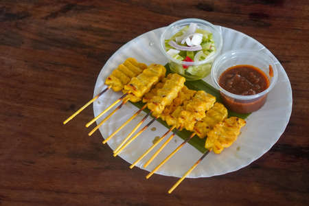 hot ten sticks of pork satay with dipping sauce, placed on a banana leaf on a paper plate, on wooden background, food, copy spaceの写真素材