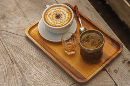 hot caramelmachiato in a white ceramic cup and honey in a glass and water in a ceramic glass and wooden spoon, place on a rectangular wooden tray,  on the wooden table background, food and drinks, copy spaceの写真素材