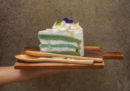 hold brown wooden chopping board, butterfly pea cake with three layer of cake on top by coconut and a butterfly pea flower, sprinkle with almond slices and two light brown wooden spoons, on cement backgroundの写真素材