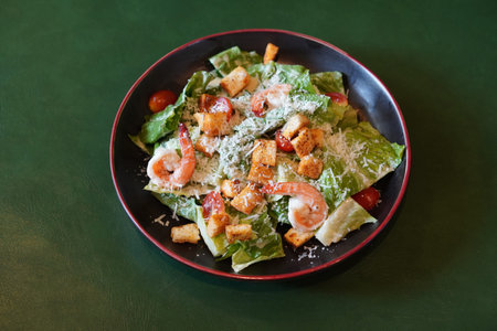 top view fresh salad with shrimp, croutons, tomatoes, and green vegetables in a ceramic black and red plate on green leather floor backgroundの写真素材