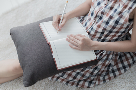 Beautiful young woman writing something in the note pad while lying on the floor at living roomの写真素材