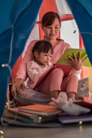 reading and family games in children's tent. asian mother and child daughter with book and flashlight before going to bedの写真素材