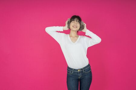Adorable asian woman in white shirt singing and having fun while listening to music using wireless earphones isolated over pink backgroundの写真素材