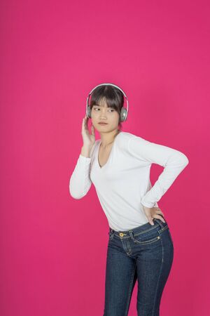 Adorable asian woman in white shirt singing and having fun while listening to music using wireless earphones isolated over pink backgroundの写真素材