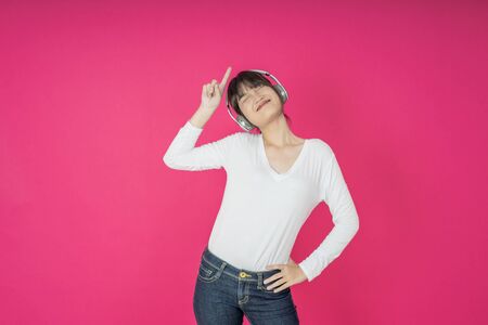 Adorable asian woman in white shirt singing and having fun while listening to music using wireless earphones isolated over pink backgroundの写真素材