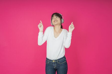 Adorable asian woman in white shirt singing and having fun while listening to music using wireless earphones isolated over pink backgroundの写真素材