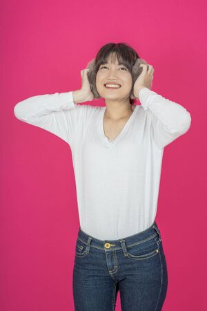 Adorable asian woman in white shirt singing and having fun while listening to music using wireless earphones isolated over pink backgroundの写真素材