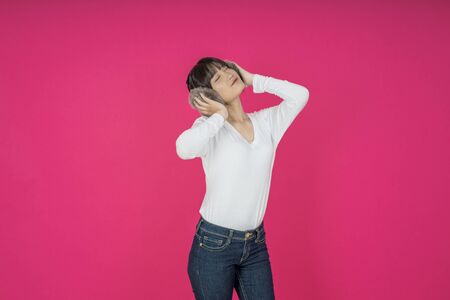 Adorable asian woman in white shirt singing and having fun while listening to music using wireless earphones isolated over pink backgroundの写真素材
