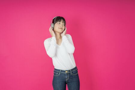 Adorable asian woman in white shirt singing and having fun while listening to music using wireless earphones isolated over pink backgroundの写真素材