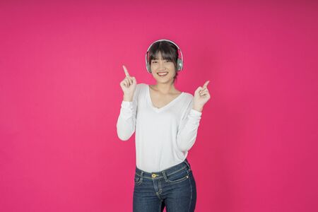 Adorable asian woman in white shirt singing and having fun while listening to music using wireless earphones isolated over pink backgroundの写真素材