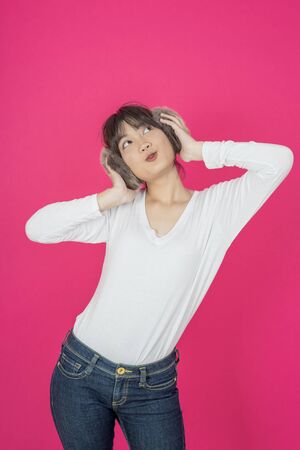 Adorable asian woman in white shirt singing and having fun while listening to music using wireless earphones isolated over pink backgroundの写真素材