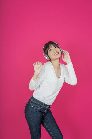 Adorable asian woman in white shirt singing and having fun while listening to music using wireless earphones isolated over pink backgroundの写真素材