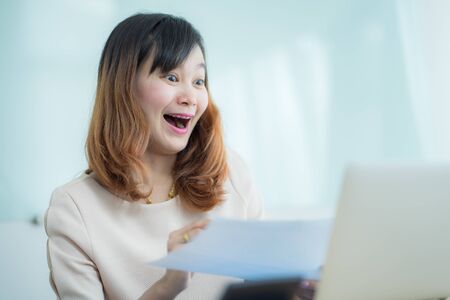 Asian businesswoman laughing with joy at workplace, gladly looking at laptop screen, feeling excited about online win, watching funny video on computer, enjoying positive good news in internetの写真素材