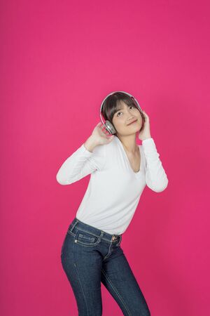 Adorable asian woman in white shirt singing and having fun while listening to music using wireless earphones isolated over pink backgroundの写真素材