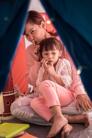 reading and family games in children's tent. asian mother and child daughter with book and flashlight before going to bedの写真素材