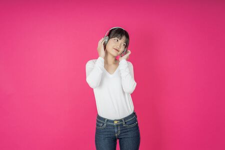 Adorable asian woman in white shirt singing and having fun while listening to music using wireless earphones isolated over pink backgroundの写真素材