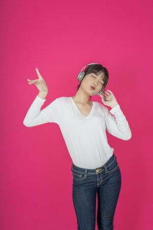 Adorable asian woman in white shirt singing and having fun while listening to music using wireless earphones isolated over pink backgroundの写真素材