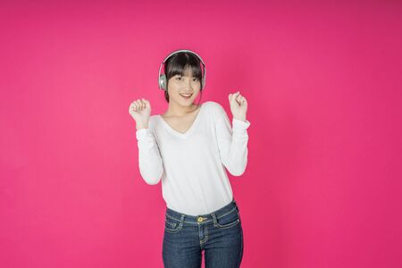 Adorable asian woman in white shirt singing and having fun while listening to music using wireless earphones isolated over pink backgroundの写真素材
